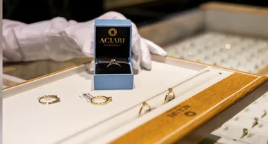 Ręka w białej rękawiczce trzyma otwarte pudełko z pierścionkiem nad tacką z biżuterią.