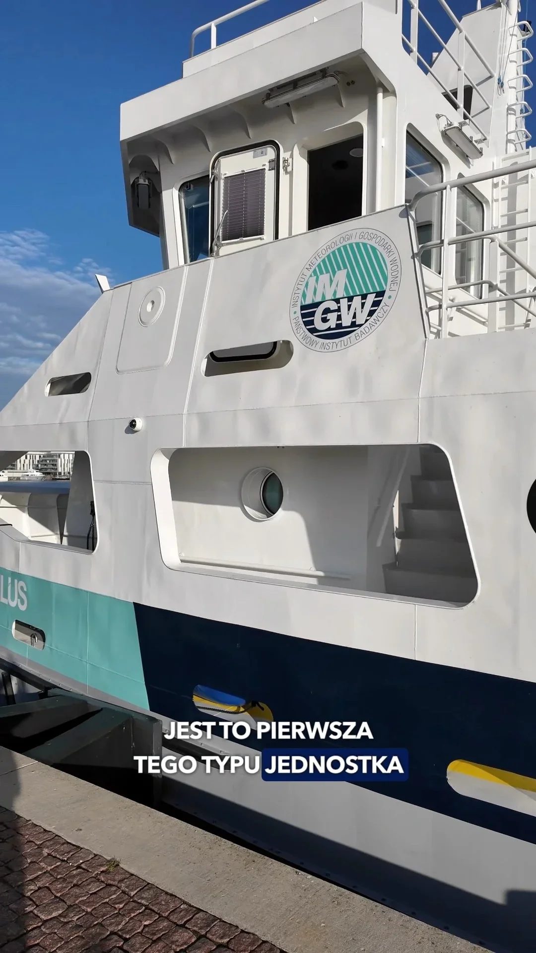 Fragment białej jednostki pływającej z logo IM GW, cumującej przy nabrzeżu w słoneczny dzień.