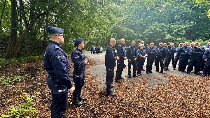 Funkcjonariusze policji w mundurach zgromadzeni na leśnej drodze podczas odprawy służbowej.