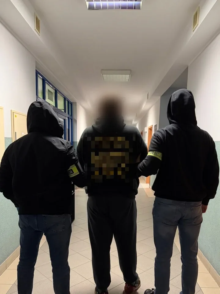 Trzej mężczyźni w czarnych bluzach z kapturami, z których dwaj boczni mają opaski na ramionach, stoją plecami do obiektywu w oświetlonym korytarzu budynku użyte