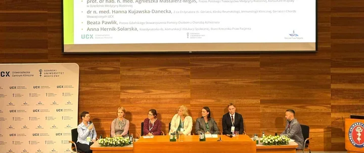 Panel dyskusyjny z sześcioma uczestnikami siedzącymi przy stole na sali konferencyjnej z drewnianą ścianą i ekranem projekcyjnym.