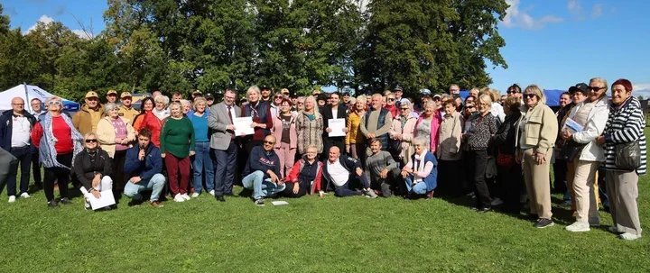 Grupa osób, głównie seniorów, pozuje do pamiątkowego zdjęcia na trawie podczas plenerowego spotkania w słoneczny dzień.