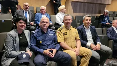 Cztery osoby, w tym funkcjonariusz policji i strażak, siedzą w pierwszym rzędzie sali audytoryjnej podczas oficjalnego spotkania lub konferencji.