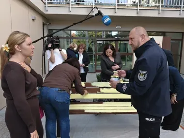 Policjant z Gdańska i kilka osób maluje ławę na żółto przed budynkiem, w tym ekipa telewizyjna nagrywa wydarzenie.