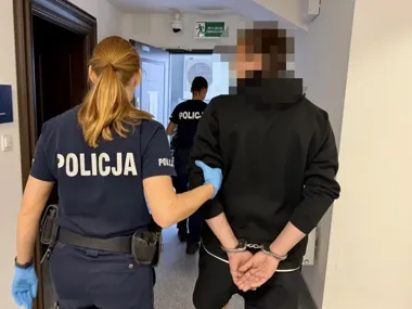Funkcjonariuszka policji w niebieskich rękawiczkach prowadzi zatrzymanego mężczyznę w kajdankach korytarzem budynku.