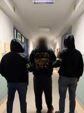 Trzej mężczyźni w czarnych bluzach z kapturami, z których dwaj boczni mają opaski na ramionach, stoją plecami do obiektywu w oświetlonym korytarzu budynku użyte