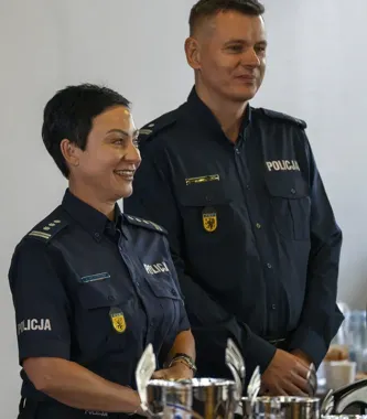 Dwie osoby w granatowych mundurach z napisem POLICJA i policyjnymi oznakami stoją przy stole zastawionym sztućcami i naczyniami.