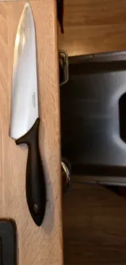 Nóż kuchenny z czarną rękojeścią leży na drewnianym blacie obok zlewu.