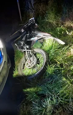 Motocykl terenowy leży na poboczu porośniętym trawą, oświetlony światłem pojazdu w nocy.