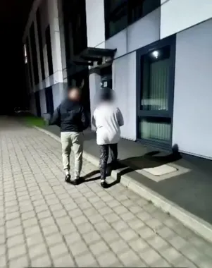 Dwie osoby o zamazanych twarzach stoją nocą na brukowanej nawierzchni przy budynku z oświetlonym wejściem.
