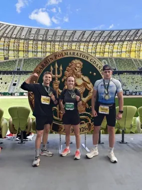Trójka biegaczy z medalami i numerami startowymi pozuje na podium przed emblematem Garmin Półmaraton Gdańsk na stadionie.