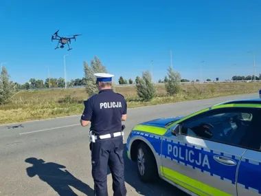 Policjant w mundurze obsługuje drona stojąc przy policyjnym radiowozie na poboczu drogi w słoneczny dzień.