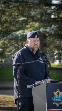 Funkcjonariusz policji w granatowym mundurze przemawia do mikrofonu przy mównicy z emblematem Policji na tle drzew iglastych.