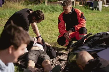 Ratownik medyczny w pomarańczowym mundurze i inna osoba udzielają pomocy mężczyźnie leżącemu na kocu na trawie obok uszkodzonego pojazdu.