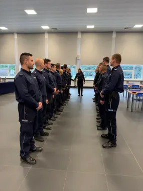 Umundurowani policjanci stoją w dwóch szeregach w sali, przed nimi znajduje się funkcjonariuszka.