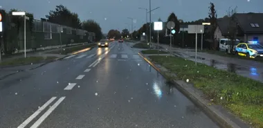 Wieczorna, mokra droga miejska z przejściem dla pieszych, pojazdami i policyjnym radiowozem z włączonymi światłami na poboczu.