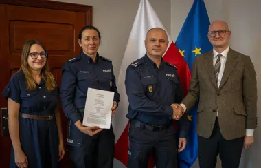 Cztery osoby pozują w pomieszczeniu z flagami Polski i UE, dwie w mundurach policji, jedna trzyma dokument, a dwie męskie osoby witały się uściskiem dłoni.