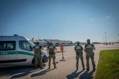 Czterech funkcjonariuszy Straży Granicznej stoi na płycie lotniska obok służbowego pojazdu, w tle widać terminale i samoloty.