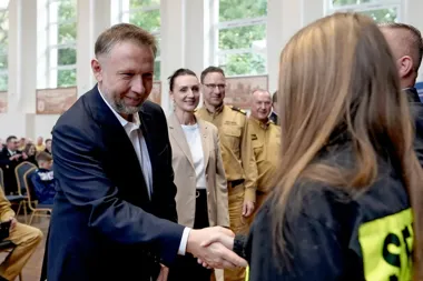 Mężczyzna w garniturze wita się uściskiem dłoni z osobą w czarnej kurtce z żółtym elementem odblaskowym, w tle stoją osoby w mundurach i eleganckim ubraniu w ja