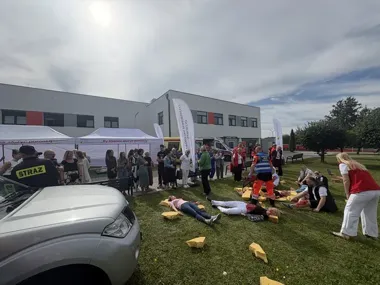 Pokaz pierwszej pomocy i działań ratowniczych na trawie przed budynkiem, z udziałem służb mundurowych, ratowników i grupy obserwatorów.