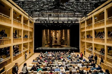 Widownia nowoczesnego teatru z drewnianymi balkonami wypełniona publicznością przed rozpoczęciem spektaklu.