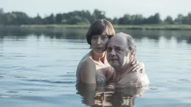 Mężczyzna i kobieta stoją w wodzie jeziora, przytuleni do siebie, z zalesionym brzegiem w tle.
