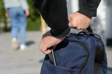 Mężczyzna w czarnej bluzie wkłada smartfon do kieszeni niebieskiego plecaka na zewnątrz, w tle przechodzi inna osoba.