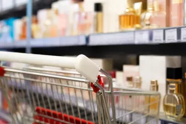 Wózek sklepowy stoi przed półkami z perfumami i kosmetykami w drogerii.