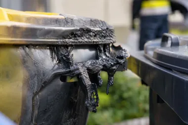 Stopiony i spalony plastikowy kosz na śmieci z żółtą pokrywą stoi obok innego pojemnika na zewnątrz.