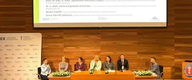 Panel dyskusyjny z sześcioma uczestnikami siedzącymi przy stole na sali konferencyjnej z drewnianą ścianą i ekranem projekcyjnym.