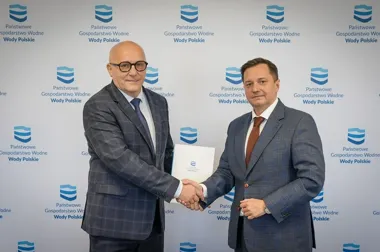 Dwaj mężczyźni w garniturach podają sobie dłonie przed ścianką z logo Państwowe Gospodarstwo Wodne Wody Polskie, jeden z nich trzyma dokument.