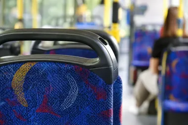 Wnętrze autobusu miejskiego z widocznymi oparciami pasażerskich siedzeń, w tle siedząca pasażerka.