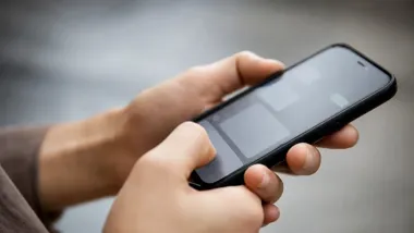 Dłonie osoby trzymające czarny smartfon z szarymi prostokątami na ekranie.