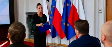 Kobieta w czarnej stylizacji z zielonymi rękawami przemawia do siedzących osób w sali z flagami Polski i Unii Europejskiej.