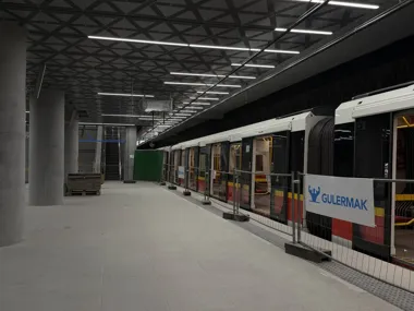 Stacja metra z nowoczesnym pociągiem stojącym przy peronie, na którym widoczne są słupy, oświetlenie i ogrodzenie z napisem wykonawcy.