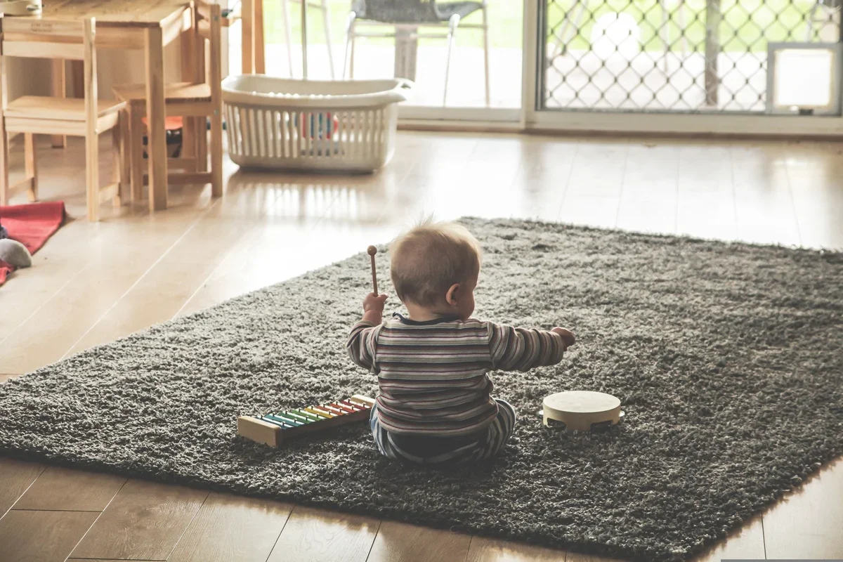 Małe dziecko siedzi tyłem na szarym dywanie w salonie i bawi się drewnianymi instrumentami muzycznymi.