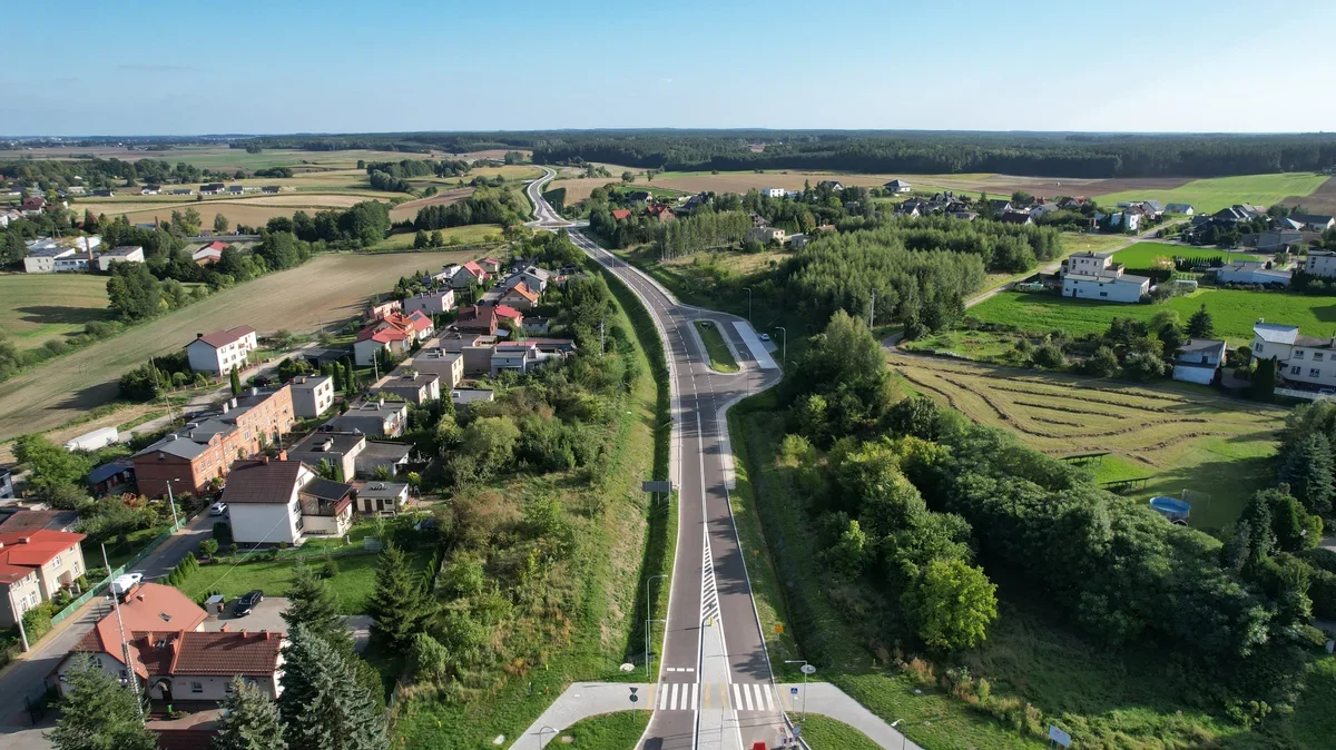 Widok z lotu ptaka na nowo wybudowaną lub wyremontowaną drogę biegnącą przez wiejską, zabudowaną okolicę otoczoną polami i lasami.